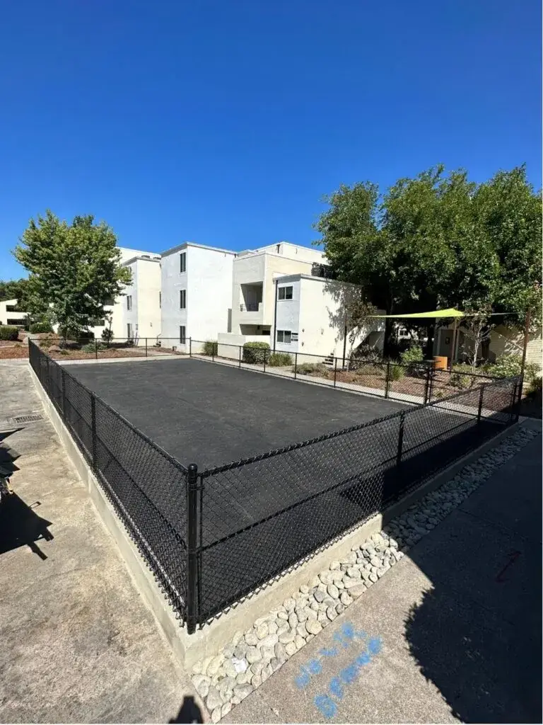 A chain-link fence surrounding a well-maintained pickleball court, with clear skies and apartment buildings in the background.