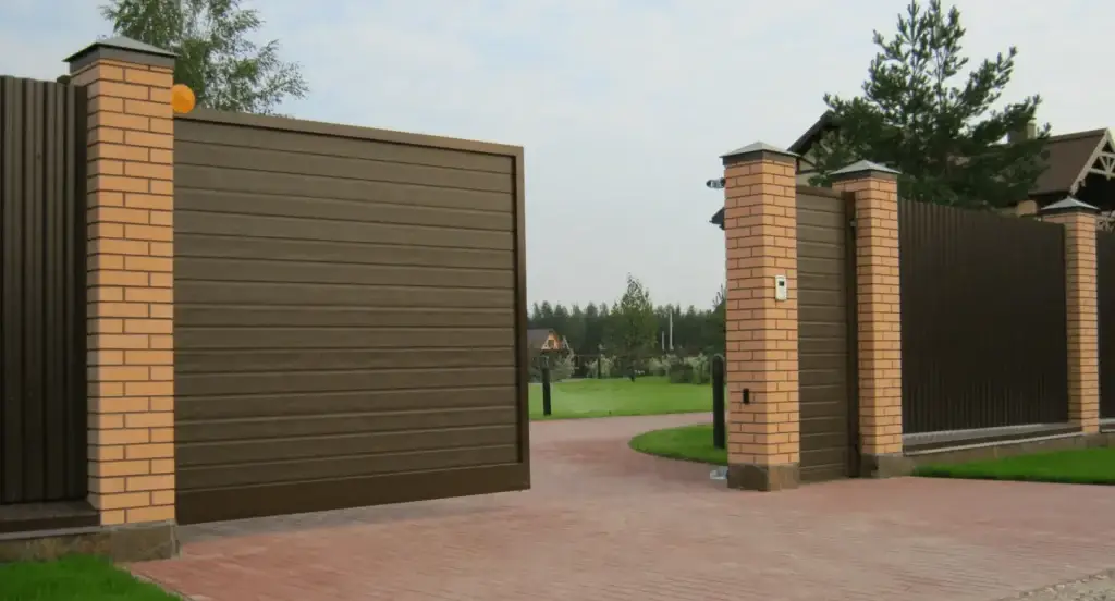 A modern metal gate with brick pillars, leading to a lush green yard and a paved driveway.