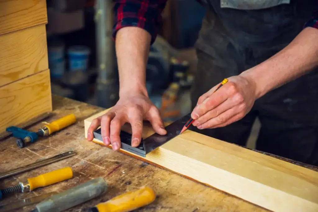 A CSLB-licensed carpenter carefully marks a wooden plank with a square tool in a woodworking workshop.